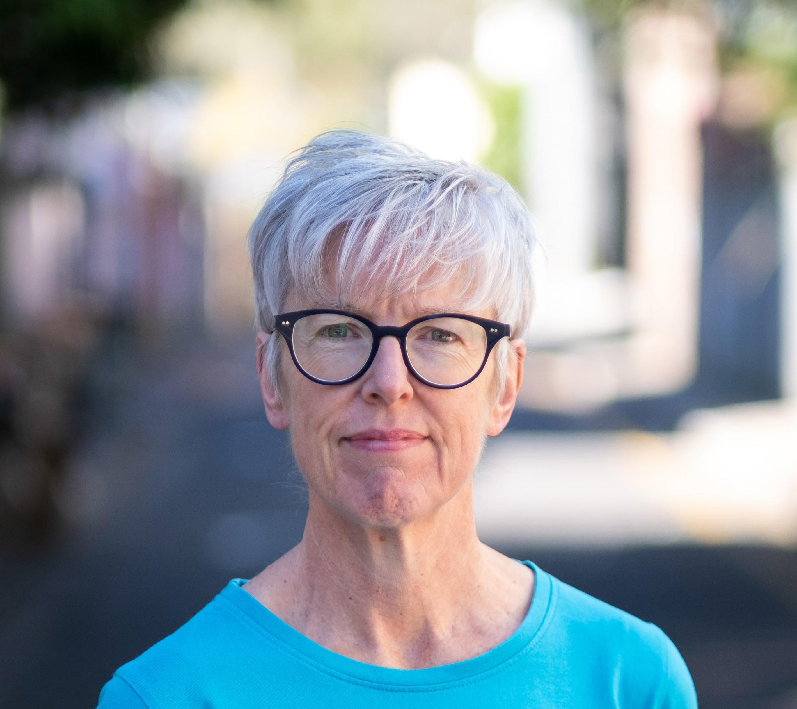 Helen Gibbons in a blue shirt, dark round glasses and a blurred background.