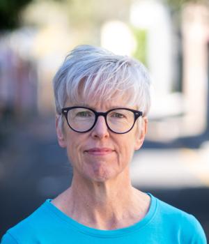 Helen Gibbons in a blue shirt, dark round glasses and a blurred background.