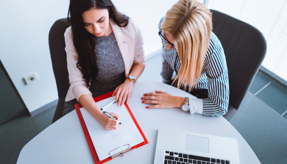 two women reviewing paperwork at a small table with laptop