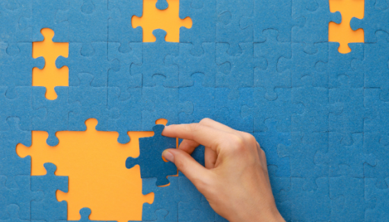 a blue puzzle on a yellow background with pieces missing and a hand putting one in to place