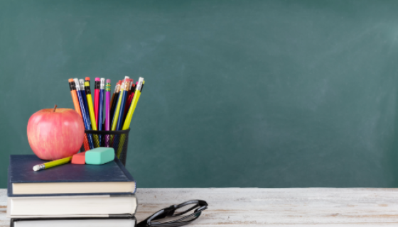 An image showing some books, pencils and an apple on a desk in a classroom