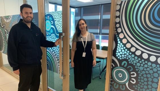 Staff members Aaron Mavrokefalos and Trish Dodd outside a new Education Standards Board meeting room featuring artwork by Aboriginal artist Allan Sumner