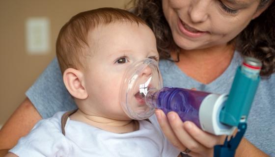 Baby being given asthma inhaler by a female carer, using a mask and spacer